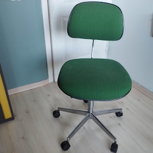 May include: A green office chair with a curved back and seat. The chair has a chrome base with five castors for mobility. The back and seat are upholstered in a textured green fabric, and the edges are trimmed with black.