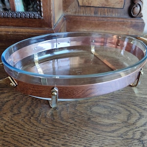 Könnte beinhalten: Eine ovale Glas-Backform mit Kupferrand und Messinggriffen. Die Form steht auf einer Holzoberfläche.