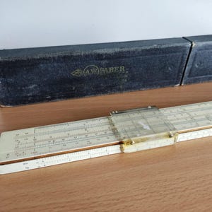 Può includere: Un regolo calcolatore vintage con un cursore in plastica trasparente e una custodia nera. Il regolo calcolatore ha segni e numeri bianchi e beige. La custodia è nera con la scritta "A.W. FABER" in lettere dorate.