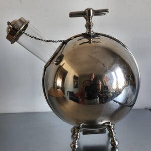 Puede incluir: Una licorera esférica de color plateado con un pico de vidrio transparente y un asa. La licorera tiene tres patas decorativas y una cadena que conecta el pico al cuerpo principal. La superficie refleja el entorno.