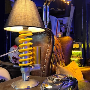 Puede incluir: Una lámpara de mesa única con una pantalla negra, una base de resorte helicoidal amarilla y plateada y un acento cromado. La lámpara está junto a un objeto cromado y un sillón de cuero. La lámpara tiene un diseño retro e industrial.