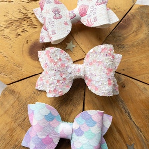 May include: Three handmade hair bows with a mermaid theme. The bows are made of white fabric with different patterns. The first bow has a pink and white seashell pattern, the second bow has pink and white glitter, and the third bow has a pink and blue mermaid scale pattern.