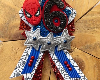 May include: A red, blue, and silver glitter birthday party pin with a Spiderman character, the number 6, and three stars. The pin is attached to a blue ribbon with a white spiderweb pattern.