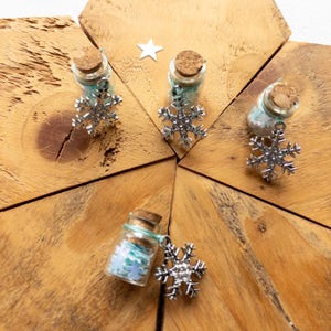 May include: Miniature glass bottles with cork stoppers, filled with blue and white confetti, and decorated with silver snowflake charms. The bottles are displayed on a wooden surface, with a small star.