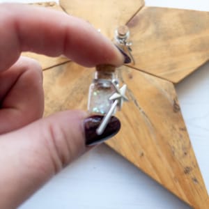 May include: A miniature glass bottle filled with glitter, held against a wooden star. The bottle has a cork stopper and a silver star charm. The wooden star has a natural wood grain finish. The bottle is filled with iridescent glitter.