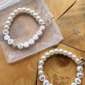 May include: Two white pearl bracelets with silver charms and the words "Big Sister" spelled out on each bracelet. The bracelets are on a wooden surface.