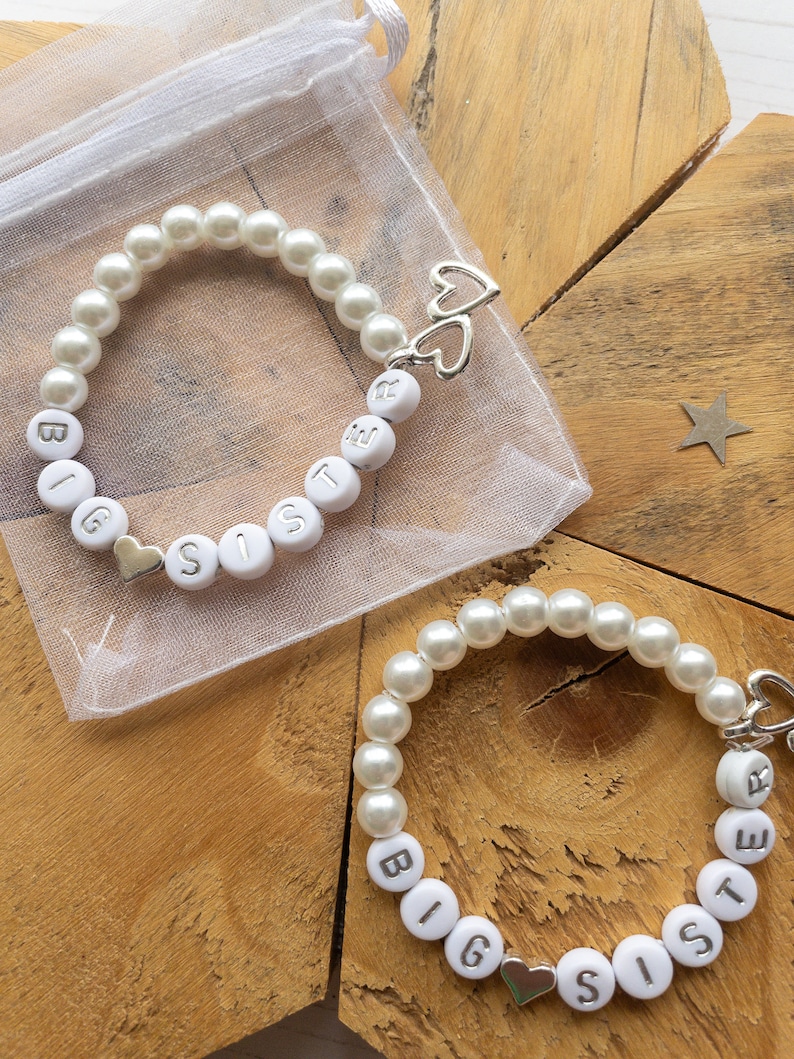 Puede incluir: Dos pulseras de perlas blancas con dijes plateados y las palabras "Big Sister" escritas en cada pulsera. Las pulseras est&aacute;n sobre una superficie de madera.