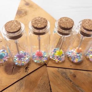 May include: Six small glass bottles with cork stoppers, each filled with colourful star-shaped confetti and a small star charm. The bottles are arranged on a wooden star-shaped surface.