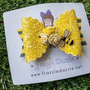 May include: A yellow glitter bow with a black and yellow striped ribbon, white flowers, and a bee charm. The bow is on a white card with the text "Frazzle Dazzle" and "www.frazzledazzle.net".