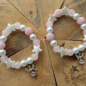 May include: Two pink and white beaded bracelets with silver star charms and a silver fairy charm. The bracelets are on a wooden surface.