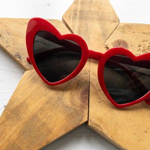 May include: Red heart-shaped sunglasses with black lenses. The sunglasses are lying on a wooden star-shaped surface.