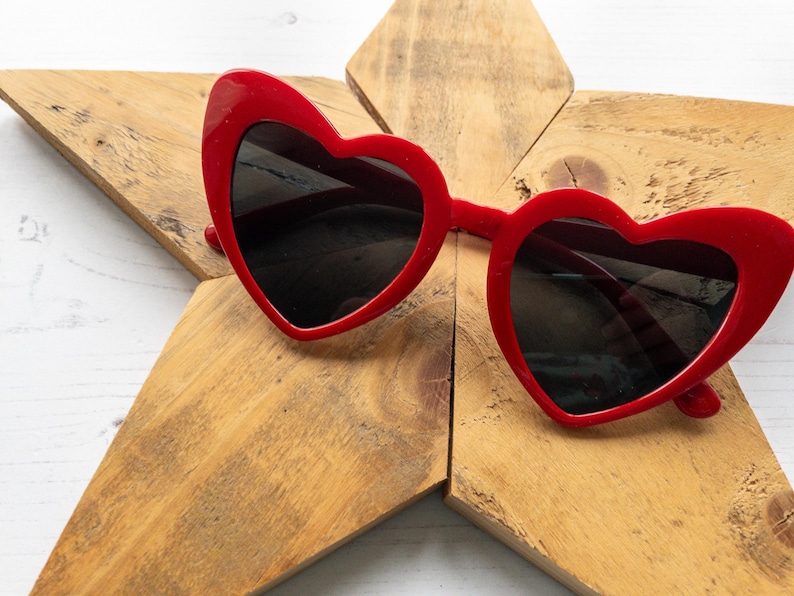 May include: Red heart-shaped sunglasses with black lenses. The sunglasses are lying on a wooden star-shaped surface.