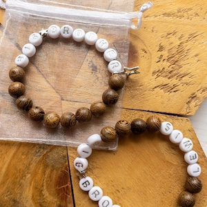 May include: Two bracelets with wooden and white letter beads spelling "BROTHER" and "SIMEON". The bracelets have a mix of dark brown and white beads. One bracelet is inside a small, sheer, white drawstring bag.