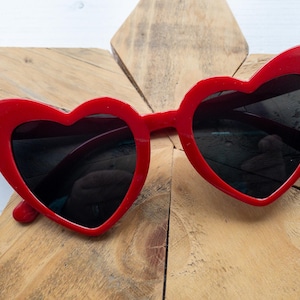 May include: Red heart-shaped sunglasses with black lenses. The sunglasses are on a wooden surface.