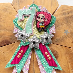 May include: A handmade birthday badge with a glittery design. The badge features a large number 7, a cartoon character, and three silver star accents. The badge has a green and pink ribbon with pearl embellishments. The background is a wooden surface.
