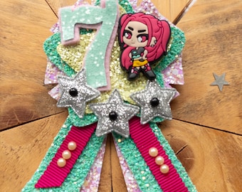 May include: A handmade birthday badge with a glittery design. The badge features a large number 7, a cartoon character, and three silver star accents. The badge has a green and pink ribbon with pearl embellishments. The background is a wooden surface.