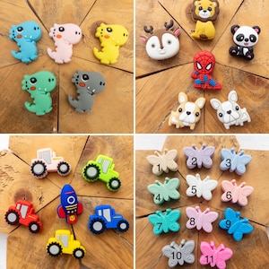 May include: A collection of silicone charms featuring various designs including dinosaurs, animals, a superhero, tractors, a rocket ship, and butterflies. The butterfly charms are numbered one through eleven in black text on their surfaces.