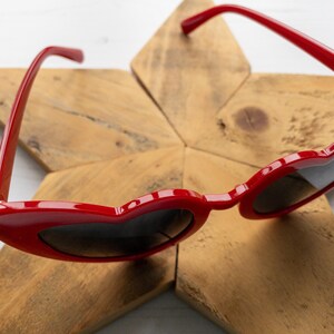 May include: Red heart-shaped sunglasses with black lenses on a wooden star-shaped surface.