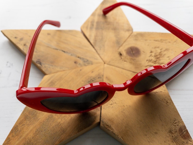 May include: Red heart-shaped sunglasses with black lenses on a wooden star-shaped surface.