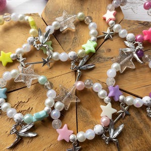 May include: Six white pearl bracelets with silver fairy charms and colourful star beads. The bracelets feature a variety of colours including pink, purple, green, blue, and yellow.