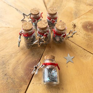 May include: Small glass bottles with cork stoppers, filled with silver star-shaped confetti. Each bottle is decorated with a red ribbon and a silver star charm. The bottles are arranged on a wooden surface.