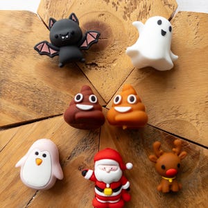 May include: A collection of seven small, colourful character charms including a black bat with pink wings, a white ghost, two brown poop emojis, a light pink penguin, a Santa Claus figure in a red suit, and a brown reindeer with a red nose.