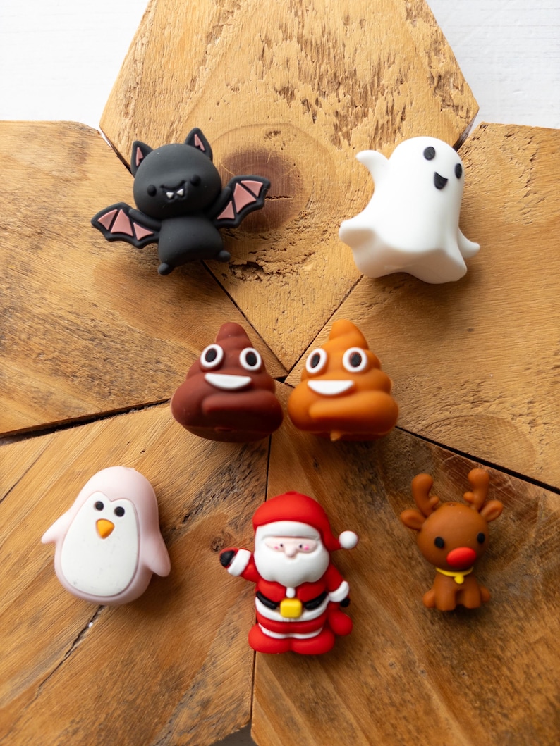 May include: A collection of seven small, colourful character charms including a black bat with pink wings, a white ghost, two brown poop emojis, a light pink penguin, a Santa Claus figure in a red suit, and a brown reindeer with a red nose.