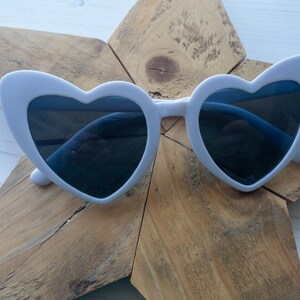 May include: White heart-shaped sunglasses with dark lenses. The sunglasses are on a wooden background.