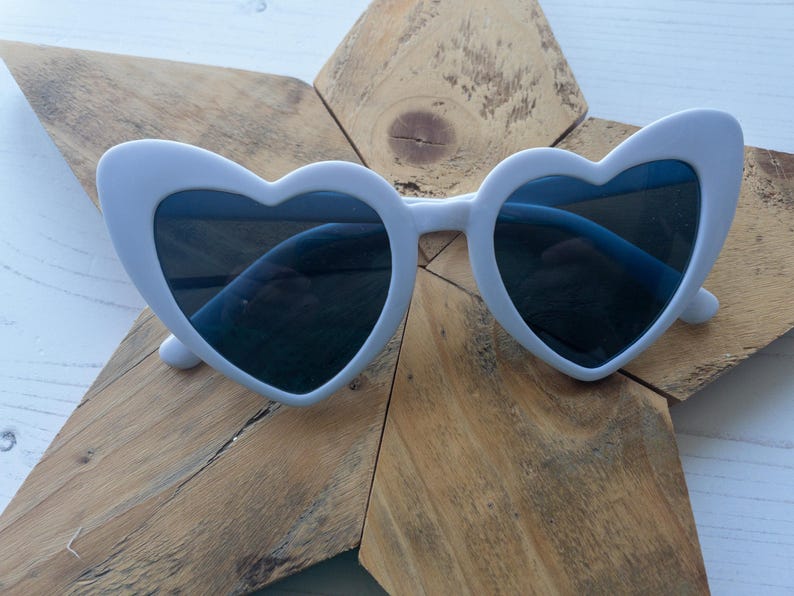 May include: White heart-shaped sunglasses with dark lenses. The sunglasses are on a wooden background.