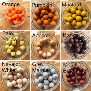 May include: A grid of nine glass bowls containing round silicone beads in various colours labelled Orange, Pumpkin, Mustard, Pale Yellow, Apricot, Brown, Navajo White, Grey Marble, and Metallic Burgundy, each with a central hole for threading.