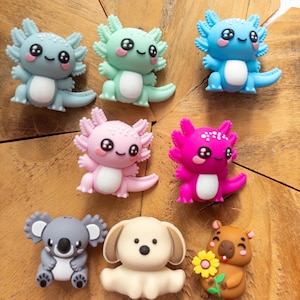 May include: A collection of eight small, colourful animal-shaped charms. The set includes five axolotls in shades of grey, mint green, blue, light pink, and magenta, alongside a grey koala, a cream-coloured puppy, and a brown capybara holding a yellow flower.