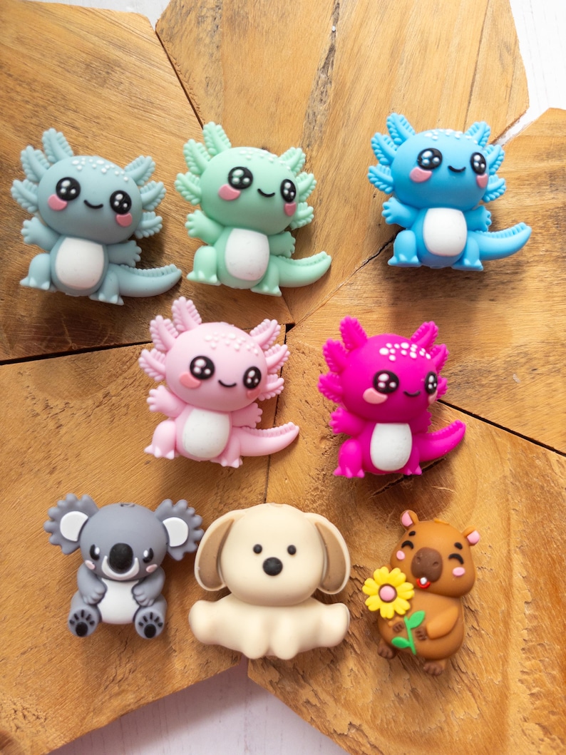 May include: A collection of eight small, colourful animal-shaped charms. The set includes five axolotls in shades of grey, mint green, blue, light pink, and magenta, alongside a grey koala, a cream-coloured puppy, and a brown capybara holding a yellow flower.