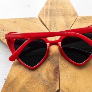May include: Red heart-shaped sunglasses with black lenses. The sunglasses are lying on a wooden surface.