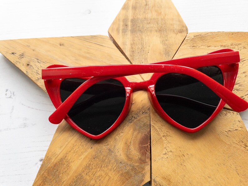May include: Red heart-shaped sunglasses with black lenses. The sunglasses are lying on a wooden surface.