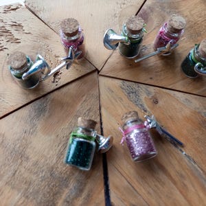 May include: Small glass bottles filled with colourful glitter, each with a cork stopper and a silver wand-shaped charm. The bottles are arranged on a wooden surface, creating a whimsical and decorative display.