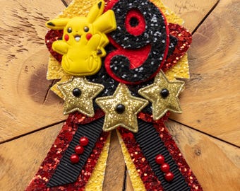 May include: A handmade badge featuring a yellow Pikachu figure, a large number 9, and three gold glitter stars. The badge has red and yellow glitter ribbon streamers and black ribbon accents. The number 9 is black with silver glitter.
