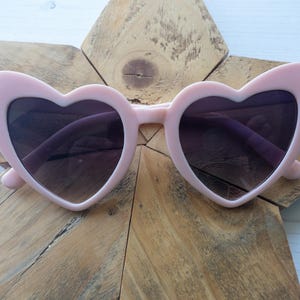 May include: Pink heart-shaped sunglasses with dark lenses. The sunglasses are on a wooden background.