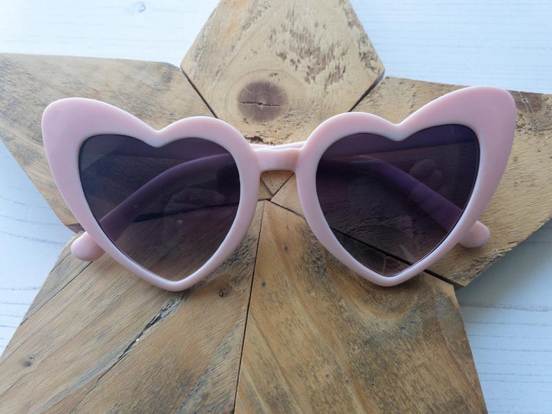 May include: Pink heart-shaped sunglasses with dark lenses. The sunglasses are on a wooden background.