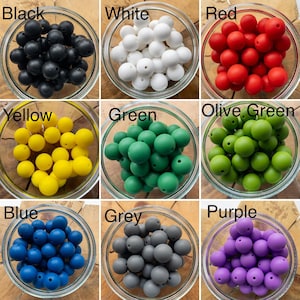 May include: A collection of nine glass bowls, each filled with round silicone beads in distinct colours labelled Black, White, Red, Yellow, Green, Olive Green, Blue, Grey, and Purple. Each bead features a central hole for threading.