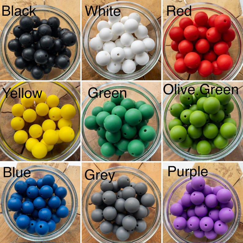 May include: A collection of nine glass bowls, each filled with round silicone beads in distinct colours labelled Black, White, Red, Yellow, Green, Olive Green, Blue, Grey, and Purple. Each bead features a central hole for threading.