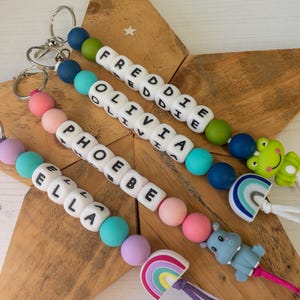 May include: Four personalised keychains featuring colourful silicone beads and white letter beads spelling the names FREDDIE, OLIVIA, PHOEBE, and ELLA. Each keychain includes a metal ring and a silicone charm shaped like a frog, rainbow, or hippo.