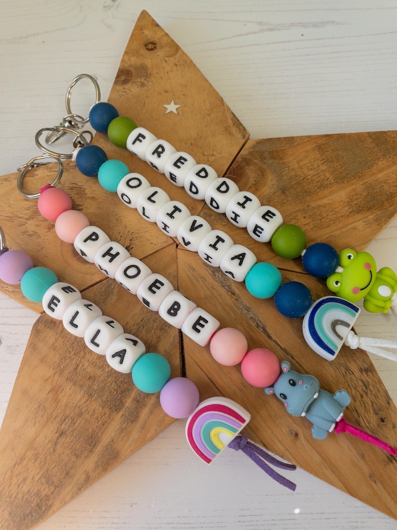 May include: Four personalised keychains featuring colourful silicone beads and white letter beads spelling the names FREDDIE, OLIVIA, PHOEBE, and ELLA. Each keychain includes a metal ring and a silicone charm shaped like a frog, rainbow, or hippo.