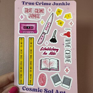 May include: A pink sticker sheet with various true crime-themed illustrations. Includes a knife, cassette tape, fingerprint, and the words "True Crime Junkie," "Allegedly," and "Establishing An Alibi." The artist's name, "Cosmic Sol Art," is also visible.