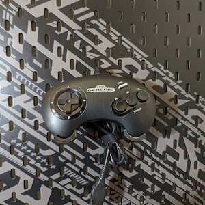 May include: A black Sega Genesis controller with a gray and black patterned background. The controller has a black cord attached to it. The controller has a start button, a mode button, and a trigger button.