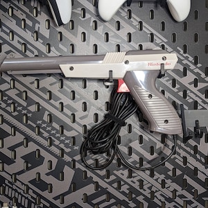 May include: A gray and white Nintendo Zapper light gun with a black cord. The gun is mounted on a pegboard wall.
