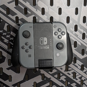 May include: A gray Nintendo Switch console with black joy-cons. The console has the Nintendo Switch logo and the text "NINTENDO SWITCH" printed on it.