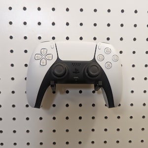 May include: A white and black video game controller with a PlayStation logo. The controller is hanging on a white pegboard.