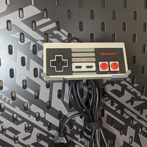 May include: A white Nintendo Entertainment System controller with a gray cord. The controller has a red and white Nintendo logo, black buttons, and a gray directional pad.
