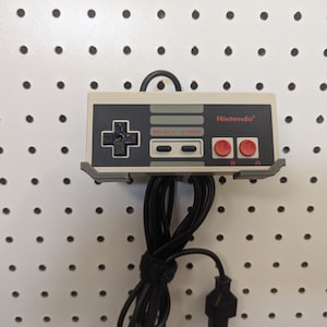 May include: A white and gray Nintendo Entertainment System controller with a black cord hanging from it. The controller is mounted on a white pegboard.