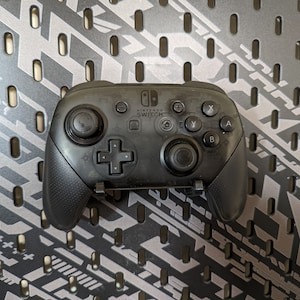 May include: A black Nintendo Switch Pro Controller with a textured grip. The controller has a D-pad, four face buttons, two analog sticks, and two shoulder buttons. The controller also has a home button, a capture button, and a power button.